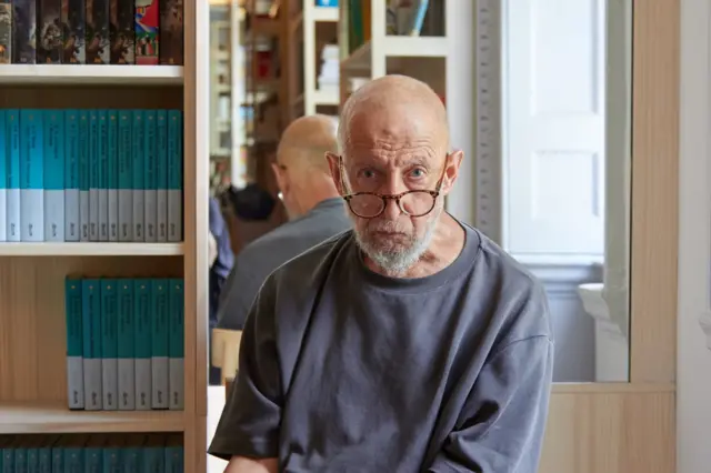 Літній сивий чоловік в окулярах сидить біля книжкових полиць. На задньому плані видно дзеркало та стелажі з книжками.