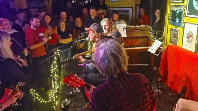 Dos músicos tocan la guitarra sentados en un escenario bajo. Uno lleva una gorra negra y el otro tiene el pelo largo y rubio. Hay luces de colores sobre un piano y una multitud los rodea.