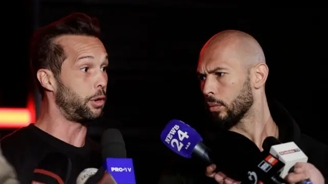 Tristan (po lewej) i Andrew Tate rozmawiają z reporterami w marcu 2025 r. Na zdjęciu widoczni są obaj bracia i szereg dłoni trzymających mikrofony. Tło jest czarne.