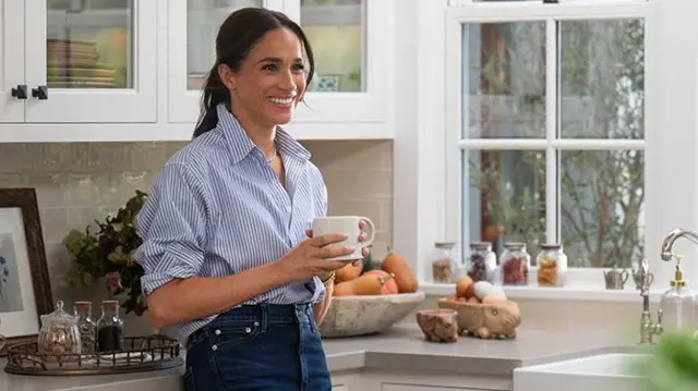 Meghan em uma cozinha com xícara nas mãos