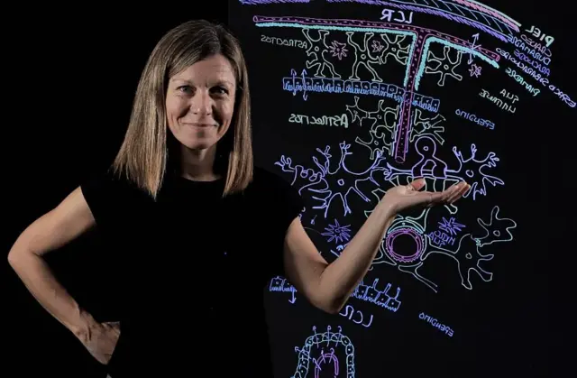 La profesora Noelia Valle, con una camiseta negra y pelo rubio en melena, frente a una pizarra con dibujos con tizas de colores sobre biología