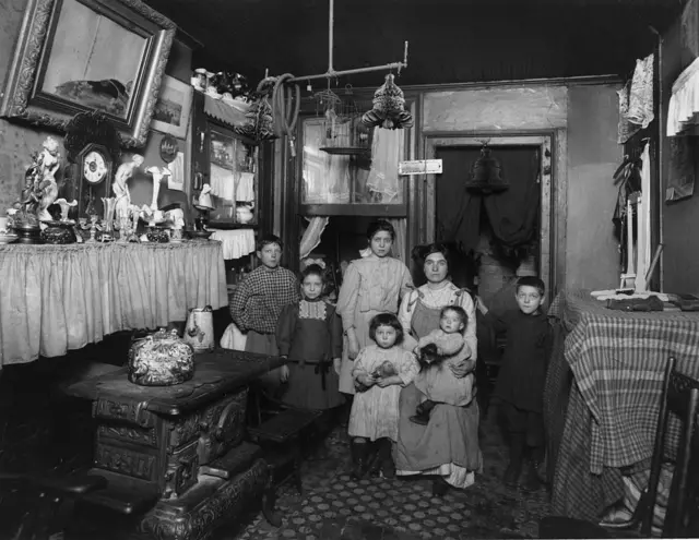 Fotografía en blanco y negro de una familia inmigrante en la ciudad de Nueva York. La madre está sentada con un bebé en el regazo.