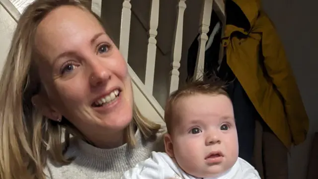 Louise sonríe a la cámara, sosteniendo a su bebé.
