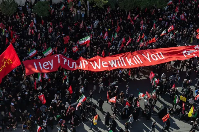 Personas en duelo participan en una procesión portando el féretro del fallecido líder supremo de Irán, el ayatolá Alí Jamenei, el 9 de julio de 2026 en Mashhad, Irán. Muestran un cartel en tela con el mensaje "Oye, Trump, te mataremos".