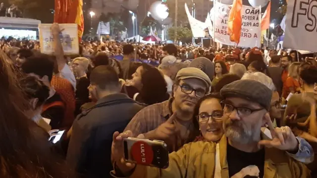 Três manifestantesmeio à multidão posam para selfie fazendo sinal com os dedos formando a letra "L"