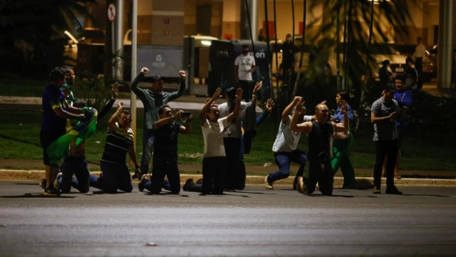 Em foto à noite, pessoas ajoelhadas, aparentemente rezando, algumas vestindo verde e amarelo e segurando bandeira do Brasil