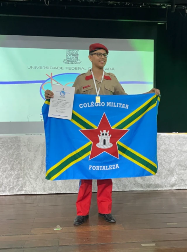 Adolescente com uniforme segurando uma bandeira onde se lê: "Colégio Militar Fortaleza"