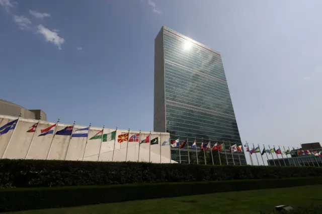المبنى عبارة عن ناطحة سحاب مليء بالنوافذ وأمامه أعلام مجموعة من الدول. الصورة التقطت نهاراً.