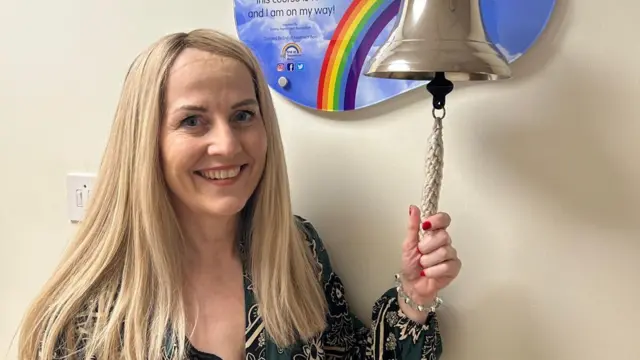 Una mujer de cabello largo y rubio aparece tocando una gran campana plateada, lo que indica su recuperación del cáncer. Lleva una blusa con estampado floral negro, blanco y verde.