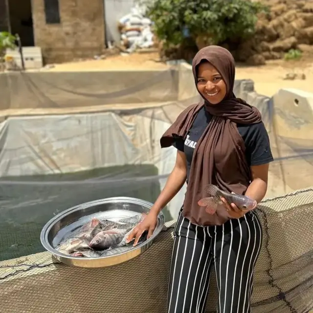 Khadija Diaw, allias Madipa à côté d'un bassin piscicole avec des poissons sur un plateau et un autre à sa main gauche.