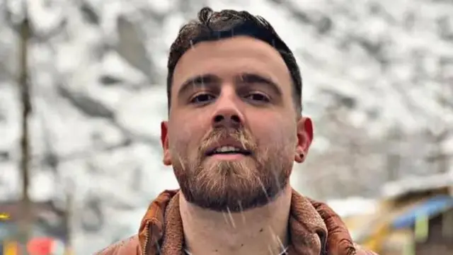 Um homem de cabelo escuro, barba e brinco na orelha olha para a câmera enquanto a neve cai ao seu redor