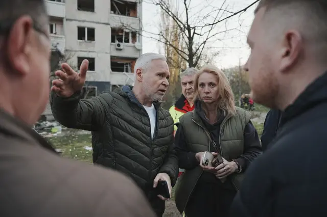 Група людей стоїть на вулиці перед пошкодженим багатоповерховим будинком, у якому вибиті вікна та видно сліди руйнувань. Терехов у центрі жестом показує рукою вбік, ніби пояснює щось про ситуацію. Інші слухають, тримаючи телефони та особисті речі. На землі видно уламки та сміття, а на задньому плані – голі дерева.