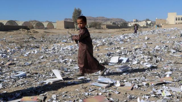 Criança caminha sobre restos de depósito de livros ecolares atingido em bombadeio no Iêmen