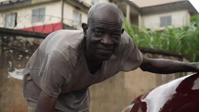 Man look straight into di camera as e put hand for car