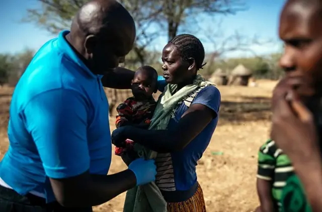 Ochieng examine des villageois. Il porte des gants, l'une des femmes tient un bébé.