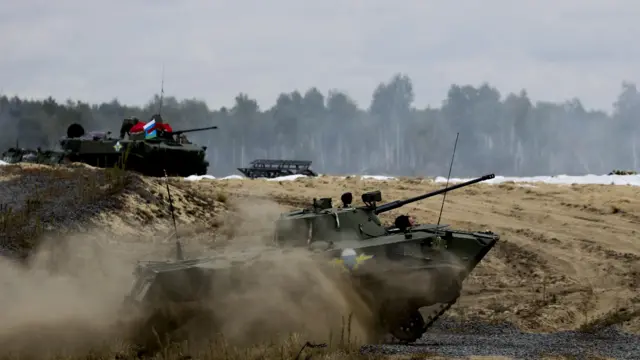 Rosyjskie i białoruskie czołgi przemierzają piaszczysty krajobraz Białorusi, wzbijając tumany kurzu. W tle widać brzozowy las. 