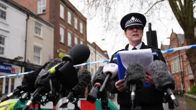 曇天の下、商店が並ぶ通りに貼られた規制線の前で、制服姿の警察幹部がクリップボードを手に、複数の取材用マイクを前に話している