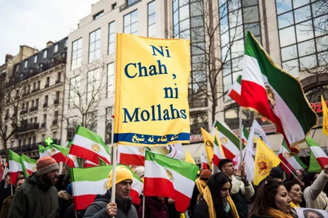 İranlı muhalifler bayrak ve pankartlarla yürüyor. Arkada apartmanlar görünüyor.