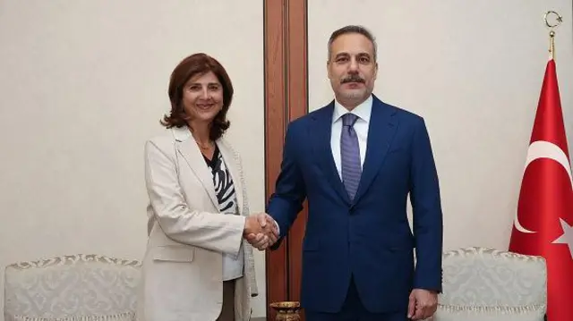 Beyaz ceketli Holguin Cuellar solda, lacivert takım elbiseli Fidan sağda el sıkışıyorlar, geride iki koltuk ve Türk bayrağı var. 