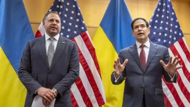 Yermak ve Rubio ayakta arkalarında ABD, Ukrayna bayrakları var ikisi de takım elbise giyinmiş, Rubio elleriyle jestler yaparak konuşuyor. 