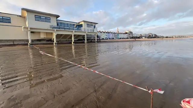 Un tramo de arena húmeda en la playa frente al edificio del Muelle de Weymouth. Se ha colocado una cinta roja y blanca para impedir que la gente camine por el lado de la playa.