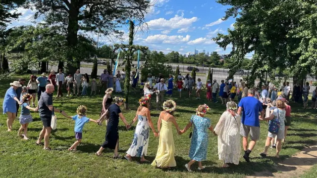 Un grupo de personas hacen un corro sujetos de la mano en un parque bajo un cielo soleado. 
