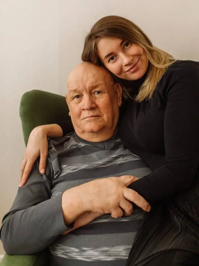 Пожилой мужчина в кресле, его обнимает молодая женщина с длинными светлыми волосами