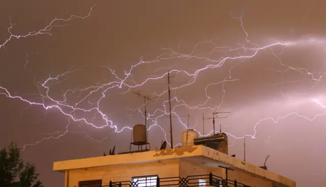 سماء مظلمة مليئة بالبرق فوق مبنى سكني يظهر من الأعلى، نافذتان مضاءتان في المبنى، وبقربه شجرة خضراء، في العاصمة الأردنية عمّان.