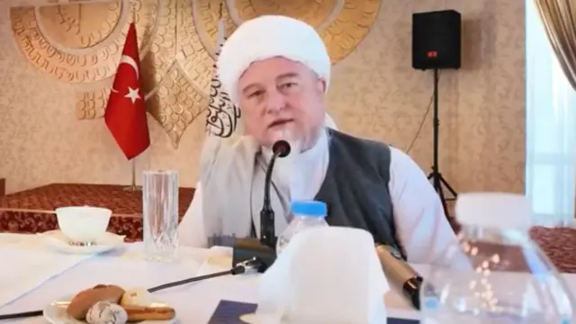 صنیعالله فرهمند، رئيس هیئت نمایندگی حکومت طالبان در ترکیه پشت میز کارش نشسته است، عمامه سفید بر سر دارد و پشت سرش پرچمهای طالبان و ترکیه قرار دارند.