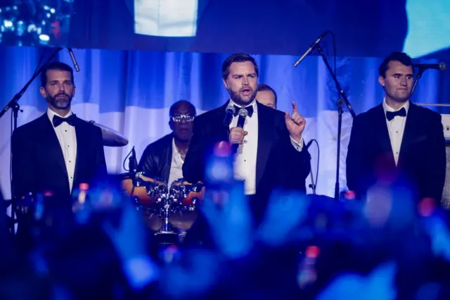 Трое мужчин в смокингах выстроились в ряд: Трамп-младший, Джей Ди Вэнс и Чарли Кирк стоят на сцене в смокингах. Вэнс (в центре) держит в руках микрофон. На заднем плане играют музыканты, на переднем плане видны руки с телефонами, фотографирующие сцену.