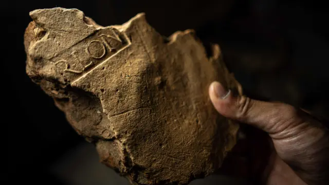 Un primer plano de un trozo roto de piedra de color marrón claro. Son claramente visibles los restos de algunas de las letras que quedaron impresas en el yeso al secarse: R-L-O-N. Significa Procurador de Londinium y muestra que el edificio del que formaba parte era un edificio civil en la ciudad en crecimiento.