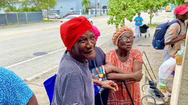 Mulheres, uma delas sorrindo para foto,sport8ng betcalçada com sacolas e carrinhos para carregar comida