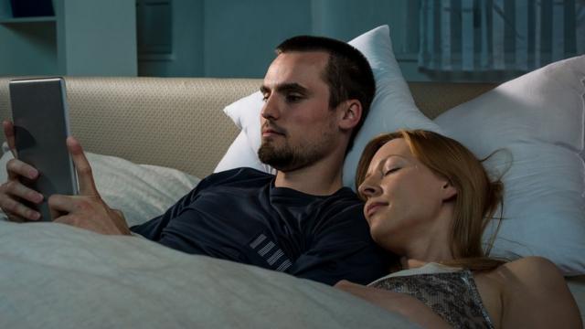 Un jeune couple allongé dans son lit ; il est sur sa tablette numérique, elle dort profondément à côté de lui.
