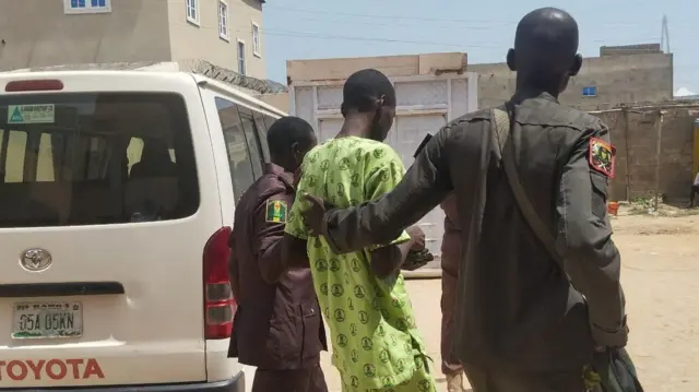 A young wey two officers dey carry go sake of im offence. Two officers dey left and right while di man dey middle