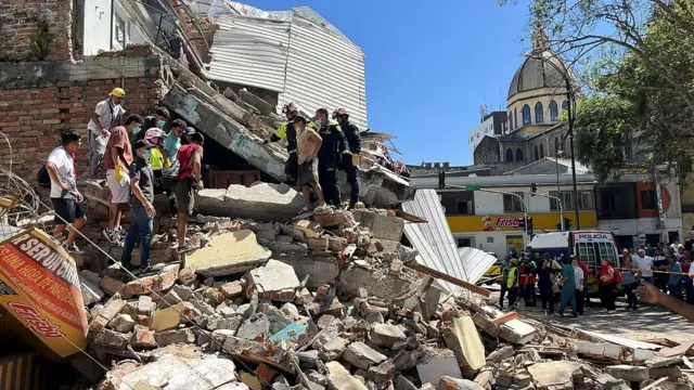 Rescatistas y personas comunes están parados sobre los escombros de un edificio colapsado mientras la gente mira desde la calle.