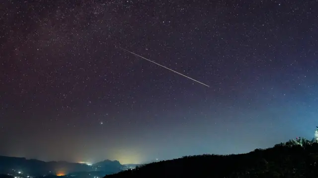 A large Perseid meteor streaks diagonally across a starry night sky above Haputale, Sri Lanka - the foreground shows dark hills with scattered lights from buildings