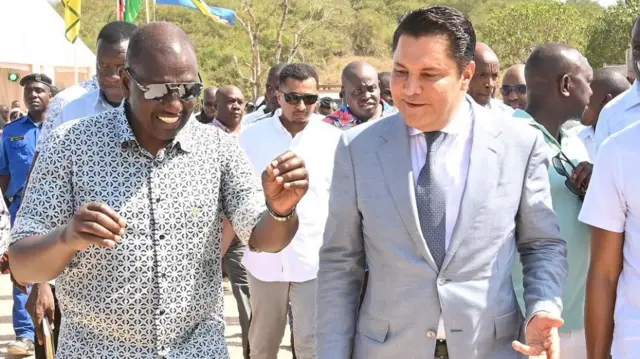 President William Ruto, wearing a checked shirt and sunglasses, walks and talks with businessman Rostam Aziz, who is wearing a grey suit