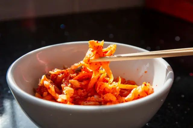 Stock image of a bowl of kimchi, with a serving held in a pair of chopsticks.
