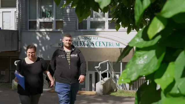 Para idzie trzymając się za ręce. Kobieta trzyma niebieski segregator. Za nimi widać budynek z napisem po węgiersku: „centrum rozrodu wspomaganego”.