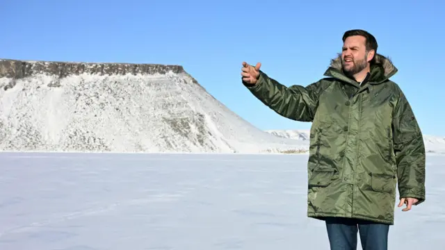 US Vice President JD Vance tours the US military’s Pituffik Space Base in Greenland on 28 March 2025. He is wearing jeans and an olive-green long puffer jacket, with a snowy field and mountains visible behind him.
