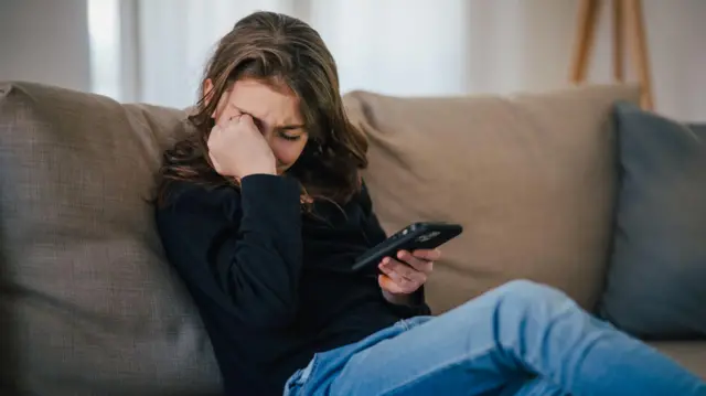 دختر نوجوانی که پیام توهین‌آمیزی را می‌خواند و گریه می‌کند؛ قربانی قلدری سایبری.