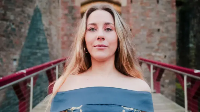 Melys Edwards - młoda kobieta o długich blond włosach, w niebieskim topie bez ramiączek - patrzy prosto w obiektyw i lekko się uśmiecha. Za nią ceglana ściana zamku i brama wejściowa.