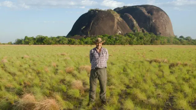 Um homem vestindo calça, camisa xadrez e um chapéu estábetnacional como jogarum campo verde. Atrás, uma imensa rocha.