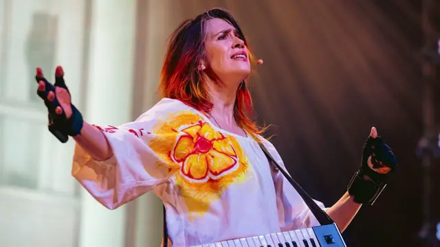 Imogen Heap występuje na scenie w Los Angeles na początku tego roku. Ma wyciągnięte ramiona, ubrana jest w biały top z pomarańczowym kwiatkiem i czarne rękawiczki.