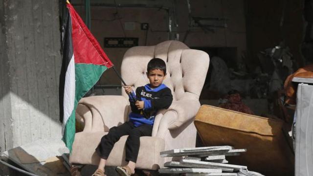 Niño sosteniendo una bandera palestina sentado en un sillón dentro de una casa derrumbada.
