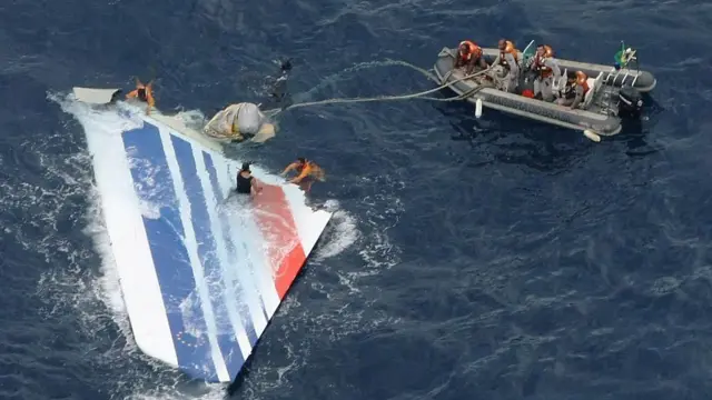 白地に赤と青の複数の線が斜めに入った旅客機の機体の一部が、海上に浮かんでいる。オレンジのライフジャケットを着た人たちが乗る灰色のゴムボートからけん引用のロープが伸び、ライフジャケットを着た人たちが水中に入り、機体を手でつかんで引いている