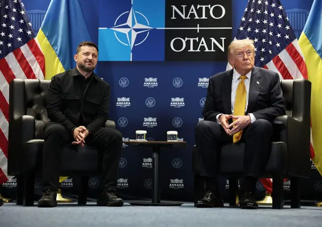 Зеленський і Трамп сидять у кріслах під час розмови на полях саміту НАТО в Анкарі. На тлі видно прапори України та США, а також логотип НАТО.