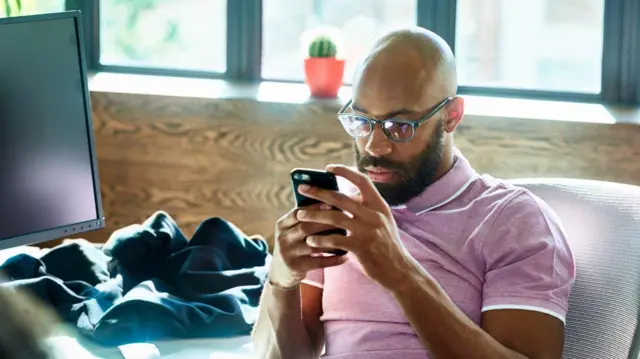 Um homem com camiseta cor de rosa olha para o celular nas mãos. 