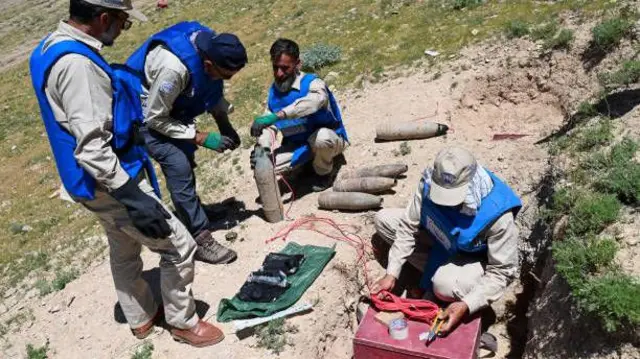 تصویر چهار مرد ماین‌پاک. آن‌ها جلیقه‌های آبی و یونیفرم خاکی به تن دارند. این مردان در حال بررسی شماری از مهمات منفجر ناشده هستند. احتمالا در حال خنثی‌سازی این مهمات اند. 