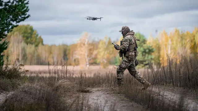 На фото військовий у камуфляжі керує дроном у польових умовах. Він стоїть на стежці серед сухої трави, тримаючи пульт управління, а перед ним у повітрі завис невеликий розвідувальний дрон. Довкола — осіння природа: дерева з жовтим і зеленим листям, сіро-блакитне небо з хмарами.